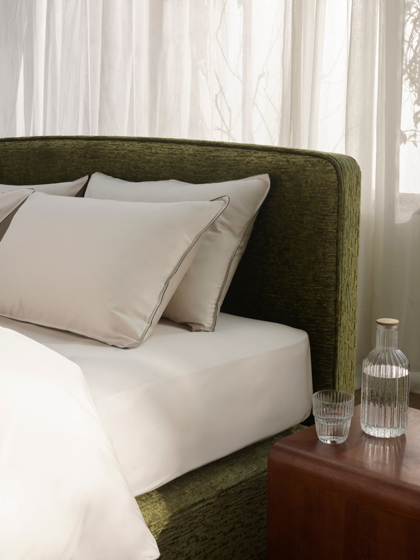 Detail of an olive chenille headboard with piped pillows and a glass carafe