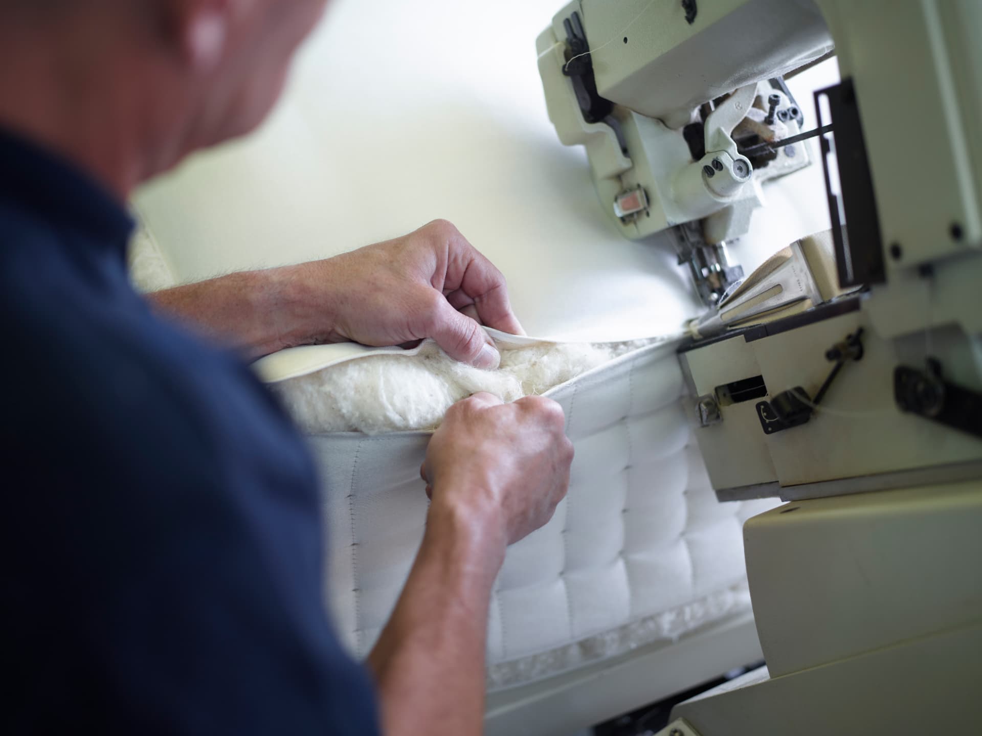 A craftsman guiding a mattress edge through a sewing machine to apply the protective tape border